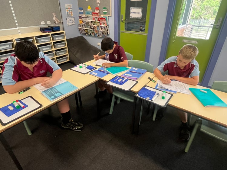 A photo of three students sitting at school desks writing in workbooks