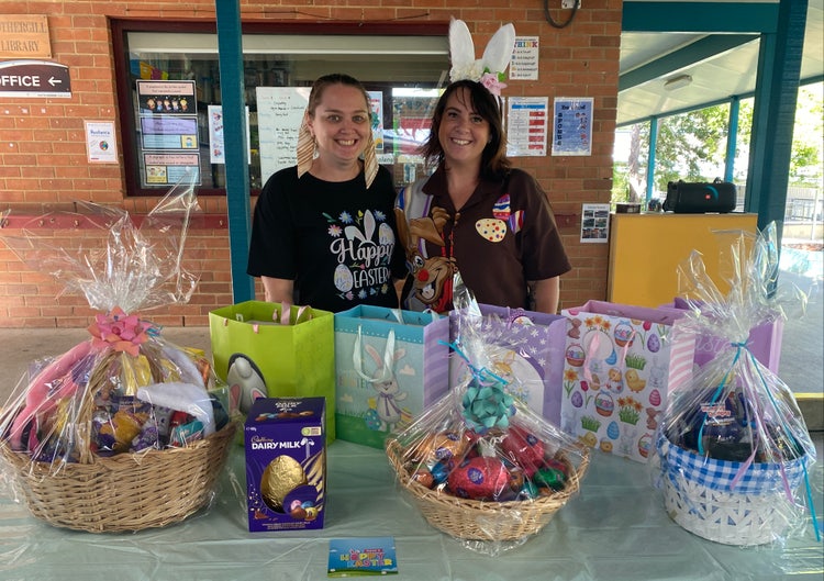 A photo of two school parents behind a table of raffle prizes baskets and gift bags
